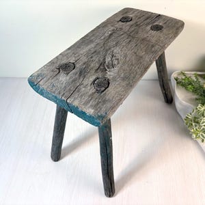May include: A small, rectangular wooden stool with a weathered, grey-brown finish. The seat features visible wood grain and four dark, circular knots. The three legs are cylindrical and dark, with traces of teal paint visible on the seat edges.