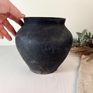 May include: A round, bulbous pot with a matte black, textured finish. The vessel features a wide, curved body that tapers towards a narrow base and a short, slightly flared rim. The surface appears weathered with subtle variations in dark tones.