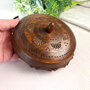 May include: A round dark brown wooden lidded box featuring intricate hand-carved geometric patterns and sunburst motifs on the lid. The lid includes a small central knob handle, and the side of the box has a serrated, scalloped edge design.