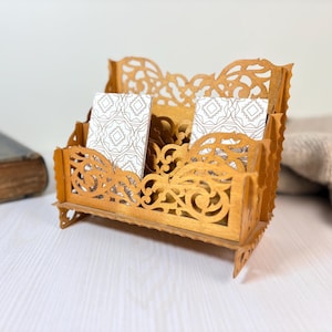 Puede incluir: Un organizador de escritorio de madera de color marrón claro con un diseño calado intrincado. El organizador contiene varias tarjetas con un patrón geométrico. El diseño presenta una estructura escalonada con recortes decorativos, ideal para la oficina o el hogar.