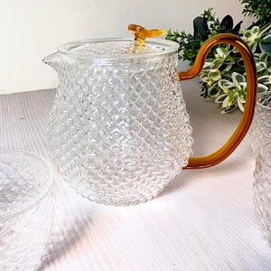 May include: A clear glass teapot featuring a textured, scale-like pattern across its body, a curved amber-coloured handle, and a matching amber-coloured knob on the lid. The teapot has a wide base and a small spout for pouring liquids.