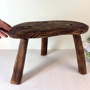 Puede incluir: Un taburete de madera de tres patas con acabado marrón oscuro. El taburete tiene un asiento redondeado con tres pequeños agujeros y está sostenido por tres patas anguladas. La veta de la madera es visible, lo que le da un aspecto rústico.