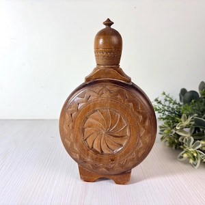 Pode incluir: Uma garrafa redonda de madeira castanha com um padrão de raios de sol esculpido no centro, rodeado por uma borda em ziguezague. A garrafa tem uma rolha de madeira a condizer com um pequeno remate no topo e acabamento liso, sobre base plana.