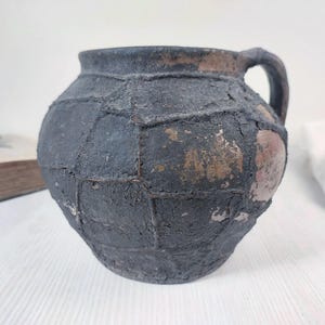 Könnte beinhalten: Ein dunkelgrauer Krug aus antiker Keramik mit Henkel. Der Krug hat eine strukturierte, patchworkartige Oberfläche mit sichtbaren Gebrauchsspuren und einigen Bereichen mit freiliegendem braunem Ton. Der Krug wird wahrscheinlich zu Dekorationszwecken verwendet.