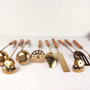 May include: A collection of vintage brass kitchen utensils with wooden handles. The set includes ladles, slotted spoons, a spatula, and a fork. The brass has a warm, golden hue, and the wooden handles are a light brown color. The utensils are arranged on a white surface.