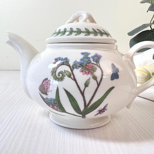 Könnte beinhalten: Weiße Keramik-Teekanne mit Deckel, Ausguss und Henkel. Die Teekanne ist mit einem Blumen- und Schmetterlingsdesign in Blau-, Rosa- und Grüntönen verziert. Der Deckel hat ein grünes Blättermuster.