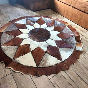 May include: A circular rug constructed from stitched panels of cowhide in shades of brown, white, and grey. The design features a central dark brown circle surrounded by alternating geometric diamond and triangular shapes, all joined by visible dark stitching.