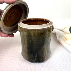 May include: A cylindrical ceramic jar with a removable lid, featuring a mottled dark green and brown glaze with visible vertical texture. The interior of the jar and the underside of the lid show a smooth, glossy brown finish with a spiral pattern.