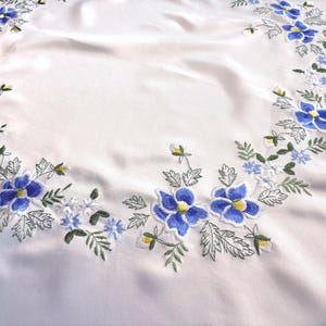 May include: White tablecloth with embroidered blue flowers and green leaves. The floral design is arranged in a circular pattern around the edge of the cloth, with yellow accents in the flower centres.