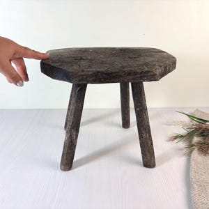 Może przedstawiać: Mały, ciemnobrązowy drewniany stołek o szorstkiej, zwietrzałej fakturze i ośmiokątnym blacie. Stołek posiada trzy solidne, cylindryczne nogi zapewniające wsparcie, prezentując naturalny, postarzany wygląd z widocznym usłojeniem drewna.