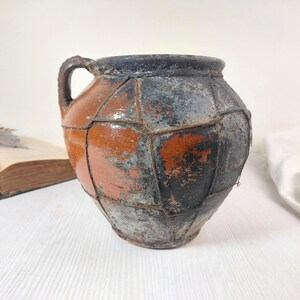 Könnte beinhalten: Ein antiker, handgefertigter Keramikkrug mit Henkel. Der Krug hat ein Patchwork-Design mit einer Mischung aus orange, schwarz und grau. Ein Beispiel für rustikale Keramik.