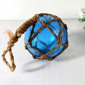 Peut inclure: Une boule en verre bleu de forme sphérique enveloppée dans un filet en corde de jute marron nouée. Le filet présente une section tressée qui s'étend de la sphère principale, formant une boucle pour suspendre l'objet. La surface est lisse.
