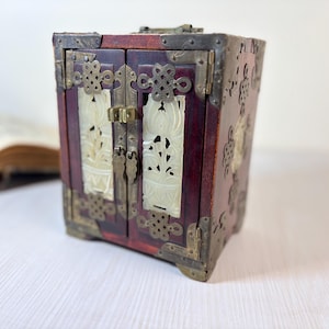 May include: A small, antique wooden box with a dark red finish and ornate brass hardware. The box features two doors with carved white panels and decorative metal accents. The box is a cube shape and has a small brass latch.