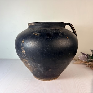 Può includere: Un grande vaso in ceramica nera arrotondato con un'ampia apertura, un collo corto e un singolo manico curvo sul lato destro. La superficie presenta una finitura opaca con segni visibili di usura, inclusi graffi e scalfitture marrone chiaro.