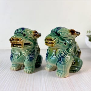 May include: Two ceramic guardian lion statues. The statues are in a seated position, with detailed features and a green, blue, and brown glaze. The lions have open mouths and are placed side by side.