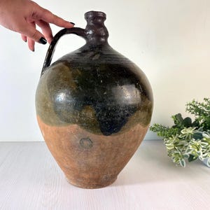 May include: A large, antique jug with a handle. The jug has a rounded body and a dark brown, glossy finish on the top half, transitioning to a lighter brown on the bottom. The jug is likely made of ceramic or earthenware.