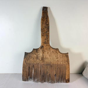 May include: A large, weathered wooden wool comb featuring a long, central handle and a wide, rectangular base. The surface displays a dark, mottled patina with visible signs of age, wear, and texture throughout the grain of the wood.