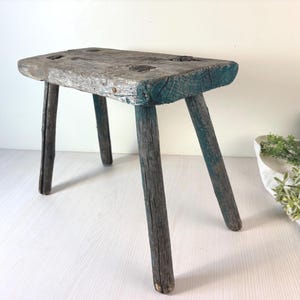May include: A small, rustic wooden stool with three legs. The stool has a rectangular seat with visible wood grain and a weathered, gray-blue finish. The legs are dark gray and angled.