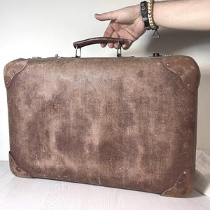 May include: A rectangular brown suitcase with a textured surface, featuring reinforced corner caps, a dark brown top handle, and metallic latches. The exterior shows signs of wear with visible scuffs and scratches across the front panel.
