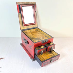 Puede incluir: Un joyero de madera rojo con tapa con bisagras y espejo incorporado. La caja tiene tres cajones pequeños y está decorada con detalles dorados y caligrafía de inspiración asiática. El interior está forrado con papel amarillo.