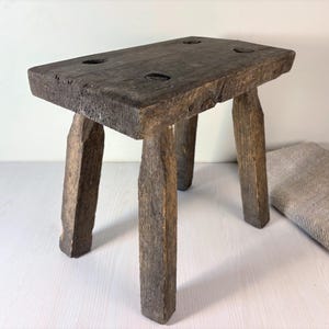 May include: A small, rectangular wooden stool with a dark, weathered finish. The stool features four thick, straight legs and a flat top surface with four visible circular joinery points at each corner, showcasing a rough, textured wood grain.