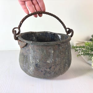 Pode incluir: Uma panela de metal de tom escuro e aspeto desgastado, com corpo arredondado e superfície texturizada irregular. Possui uma pega grossa de metal torcido fixada a dois anéis na borda, apresentando sinais de oxidação e envelhecimento.