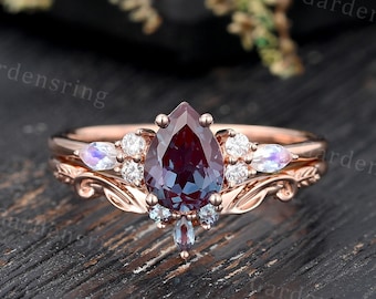 May include: Rose gold engagement ring with a pear-shaped, deep purple gemstone as the centerpiece. The ring is accented with smaller white and iridescent stones, and features a delicate, ornate band design. The ring is set on a dark wooden surface.