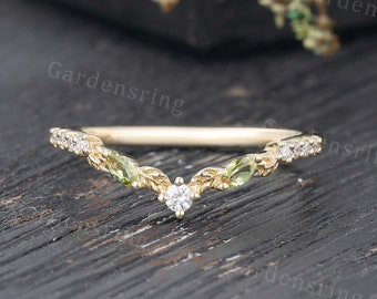 May include: A delicate gold wedding band with a V-shaped design. The band is adorned with small, clear diamonds and green gemstones. The band is set on a wooden surface.