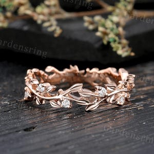 Könnte beinhalten: Roségoldener Ring mit einem Blatt- und Rankenmuster, verziert mit kleinen, klaren Edelsteinen. Der Ring ist vor einem dunklen, strukturierten Hintergrund platziert, der die Details hervorhebt.