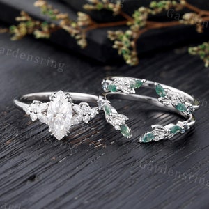 May include: A pear-shaped diamond engagement ring with a delicate band and a floral design featuring small diamonds and green gemstones. The ring is paired with a matching wedding band with a similar floral design.
