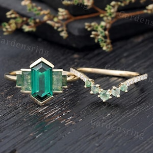 Op de afbeelding: Een gouden verlovingsring met een grote rechthoekige smaragdgroene middelste steen en kleinere smaragdgroene stenen op de band. De bijpassende trouwring is een delicate gouden band met een rij kleine smaragdgroene stenen in een v-vorm.