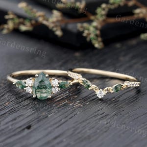 May include: A pear-shaped green agate engagement ring with a delicate band of small diamonds and green agate stones. The ring is paired with a matching wedding band featuring a V-shaped design with small diamonds and green agate stones.