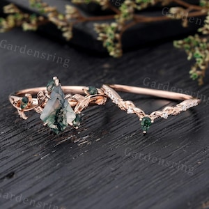 May include: A rose gold engagement ring set with a green moss agate centre stone and a matching band with green gemstones and small diamonds. The ring is set on a dark wooden surface.