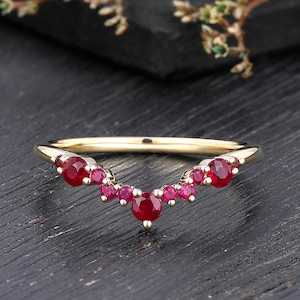 Könnte beinhalten: Ein V-förmiger Goldring mit einer Reihe von runden, facettierten roten Rubinen entlang der Ringschiene. Die Steine variieren in der Größe, wobei der größte Rubin in der Mitte des Chevron-Designs sitzt, flankiert von kleineren Rubinen.