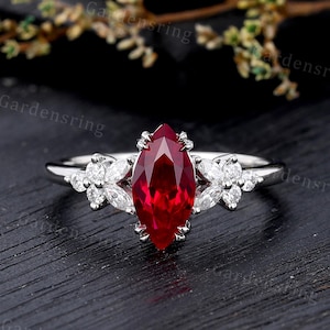 May include: A silver-coloured ring featuring a central marquise-cut red gemstone held by prongs. The band is accented on both sides by a cluster of small, clear, marquise and round-cut stones arranged in a floral-like pattern along the metal shank.