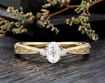 May include: A gold engagement ring with an oval-cut center stone and two smaller side stones. The band features an ornate, engraved design. The ring is set against a dark wooden surface.