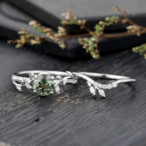 May include: A two-piece ring set. The engagement ring features a round, green gemstone and leaf accents. The wedding band is curved and adorned with small, clear gemstones. Both rings are silver-colored and set against a dark wooden surface.