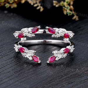 Pode incluir: Um anel aberto em metal prateado com um design inspirado em folhas. A banda é adornada com rubis vermelhos de corte marquise e pequenas pedras brancas redondas cravadas ao longo das folhas metálicas, criando uma estrutura simétrica.