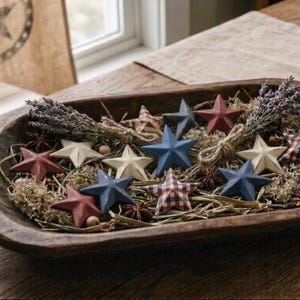 May include: A rustic wooden bowl filled with decorative stars in red, white, and blue, along with dried lavender and greenery. The stars vary in size and pattern, creating a charming, patriotic display. A burlap banner with a star design is in the background.