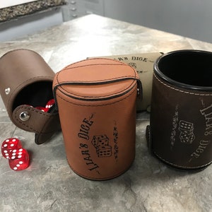 May include: Three brown leather dice cups with the text "Liar's Dice" printed on them. The cups are closed and sitting on a light brown surface.