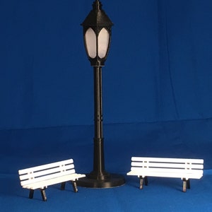 Peut inclure: Un lampadaire noir avec un abat-jour en verre blanc se trouve entre deux bancs en bois blanc sur un fond bleu.