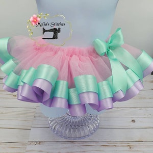 May include: A pink tulle tutu with multiple layers of lavender and mint green ribbon. The tutu has a bow on the side and is displayed on a white wooden surface. "Malia's Stitches" is written on the top of the tutu.