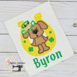 May include: A brown dog wearing a green and gold hat sits inside a yellow horseshoe with green shamrocks. The name "Byron" is embroidered in green below the horseshoe.