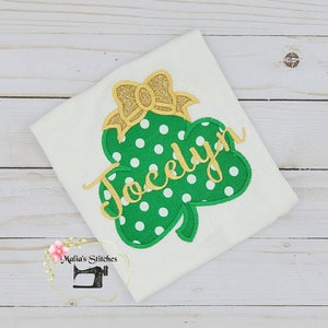 May include: A white t-shirt with a green shamrock with white polka dots and a gold bow on top. The name "Jocelyn" is written in gold cursive lettering on the shamrock.