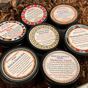 Könnte beinhalten: Sechs runde schwarze Tiegel mit Medicine Grizzly Herbs Balsamen und Salben mit bunten Etiketten. Die Etiketten enthalten Texte wie Refreshing Feet, Respiratory & Chest Balm, Warrior Women's Hands, Diabetic Balm, Working Man's Hands, Joint & Muscle Rub und Medicine Touch.
