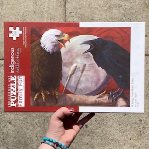 May include: A 500-piece jigsaw puzzle box featuring a painting of a bald eagle and a raven perched on a branch with a drum in the background. The puzzle is part of the Indigenous Art Collection: Family Fun by CAP. The title of the painting is "A Song From Both Sides" by Jean Taylor.