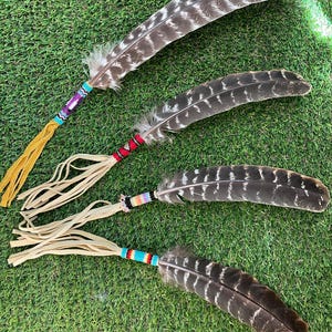 May include: Four large, brown and white striped feathers, each featuring a handle wrapped in colorful seed beads and finished with long, light tan leather fringe tassels. The beads display geometric patterns in shades of blue, purple, red, and white.