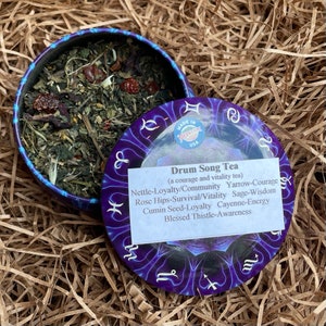 Puede incluir: Una lata redonda azul abierta con té de hojas sueltas. La tapa, decorada con símbolos del zodiaco, muestra el texto "Drum Song Tea" y una lista de ingredientes. La etiqueta también dice "Made in Montana, USA."