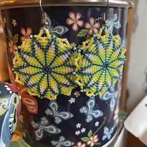 May include: A pair of circular beaded earrings featuring a radial pattern in shades of yellow, white, and dark green. The intricate beadwork creates a sunburst design with a central dark green point, suspended from silver-toned metal hooks.