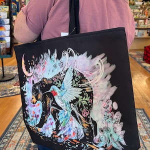 May include: A black tote bag featuring a colorful graphic print of a black bear and a hummingbird. The artwork includes swirling pastel accents, floral patterns, and a crescent moon shape, all set against the solid black fabric of the bag.